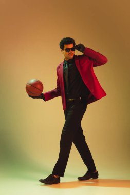 Kırmızı ceketli, yüksek moda bir adam kahverengi arka planda basketbol oynuyor.