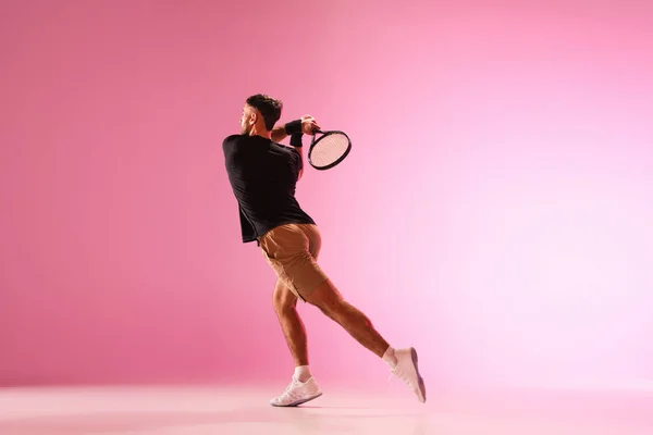 Joven hombre caucásico jugando al tenis aislado sobre fondo de estudio rosa, acción y concepto de movimiento — Foto de Stock
