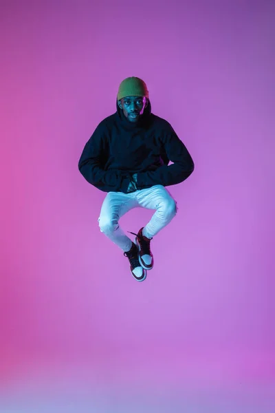 Hombre joven y elegante en traje de estilo urbano moderno aislado sobre fondo degradado en luz de neón. modelo de moda afroamericana en el libro de la mirada, músico que realiza. — Foto de Stock