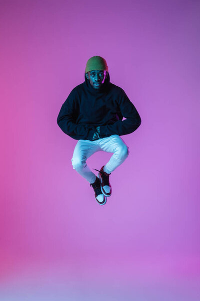 Dancer. Young stylish man in modern street style outfit isolated on gradient studio background in neon light. Full length. African-american fashionable model in look book, musician performing.