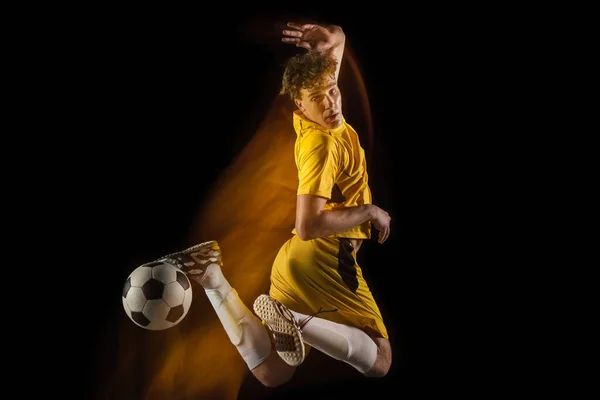 Young caucasian male football or soccer player kicking ball for the goal in mixed light on dark background. Concept of healthy lifestyle, professional sport, hobby.