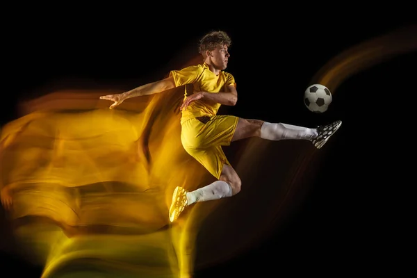 Jovem caucasiano de futebol masculino ou jogador de futebol chutando bola para o gol em luz mista sobre fundo escuro. Conceito de estilo de vida saudável, esporte profissional, hobby. — Fotografia de Stock
