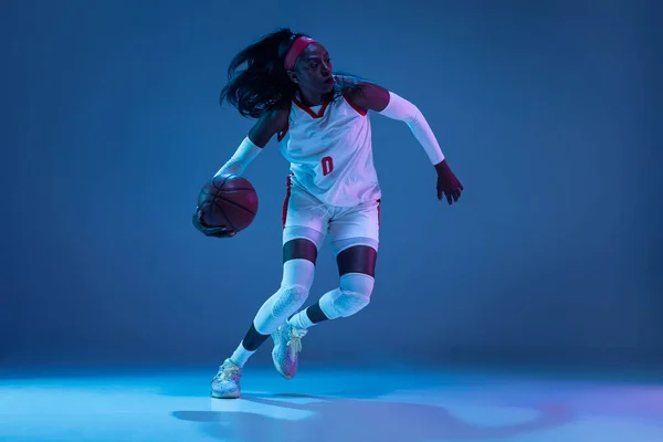 Bella giocatrice di basket afro-americana in movimento e in azione in luce al neon su sfondo blu. Concetto di stile di vita sano, sport professionale, hobby. — Foto Stock