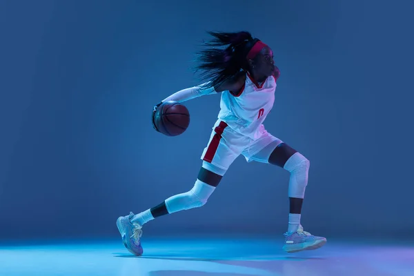 Bella giocatrice di basket afro-americana in movimento e in azione in luce al neon su sfondo blu. Concetto di stile di vita sano, sport professionale, hobby. — Foto Stock