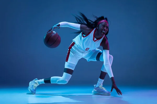 Hermosa jugadora de baloncesto afroamericana en movimiento y acción en luz de neón sobre fondo azul. Concepto de estilo de vida saludable, deporte profesional, hobby. — Foto de Stock