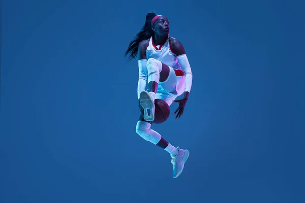 Piękna afrykańsko-amerykańska koszykarka w ruchu i akcja w neonowym świetle na niebieskim tle. Pojęcie zdrowego stylu życia, sportu zawodowego, hobby. — Zdjęcie stockowe