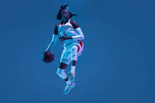 Beautiful african-american female basketball player in motion and action in neon light on blue background. Concept of healthy lifestyle, professional sport, hobby. — Stock Photo, Image