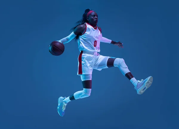 Belle joueuse afro-américaine de basket-ball en mouvement et en action au néon sur fond bleu. Concept de mode de vie sain, sport professionnel, passe-temps. — Photo
