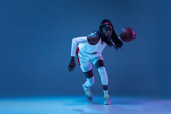 Bela jogadora de basquete afro-americana em movimento e ação em luz de néon sobre fundo azul. Conceito de estilo de vida saudável, esporte profissional, hobby. — Fotografia de Stock