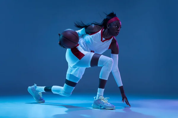 Belle joueuse afro-américaine de basket-ball en mouvement et en action au néon sur fond bleu. Concept de mode de vie sain, sport professionnel, passe-temps. — Photo