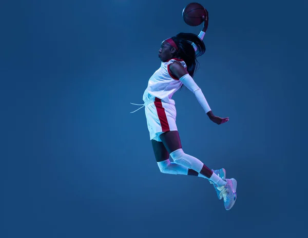 Hermosa jugadora de baloncesto afroamericana en movimiento y acción en luz de neón sobre fondo azul. Concepto de estilo de vida saludable, deporte profesional, hobby. —  Fotos de Stock