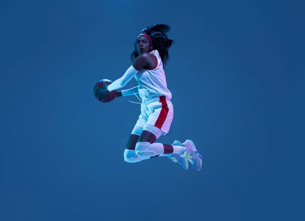 Bela jogadora de basquete afro-americana em movimento e ação em luz de néon sobre fundo azul. Conceito de estilo de vida saudável, esporte profissional, hobby. — Fotografia de Stock