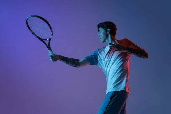 Mladý běloch hraje tenis izolovaný na fialově modrém pozadí studia v neonu, akční a pohybové koncepci — Stock fotografie