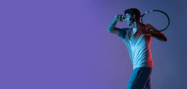 Молодий кавказький чоловік грає в теніс ізольований на фіолетово-блакитному студійному фоні в неоні, дії і концепції руху — стокове фото