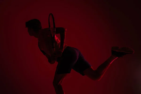Jovem caucasiano jogando tênis isolado no fundo do estúdio vermelho em neon, ação e conceito de movimento — Fotografia de Stock