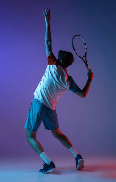 Mladý běloch hraje tenis izolovaný na fialově modrém pozadí studia v neonu, akční a pohybové koncepci — Stock fotografie