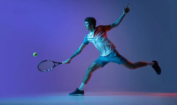Jovem caucasiano jogando tênis isolado no fundo do estúdio roxo-azul em neon, ação e conceito de movimento — Fotografia de Stock