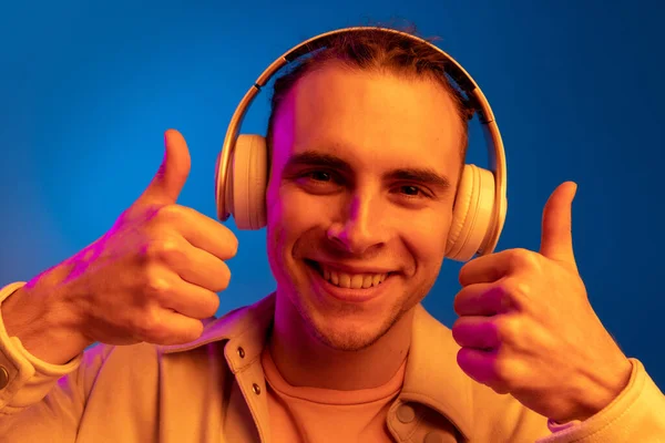 Hombre joven caucásico en auriculares aislados en fondo estudio azul en neón. Concepto de emociones humanas, expresión facial. — Foto de Stock