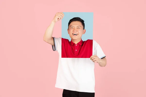 One Asian young man holding his portraits isolated on pink studio background. Concept of human emotions. — Stockfoto