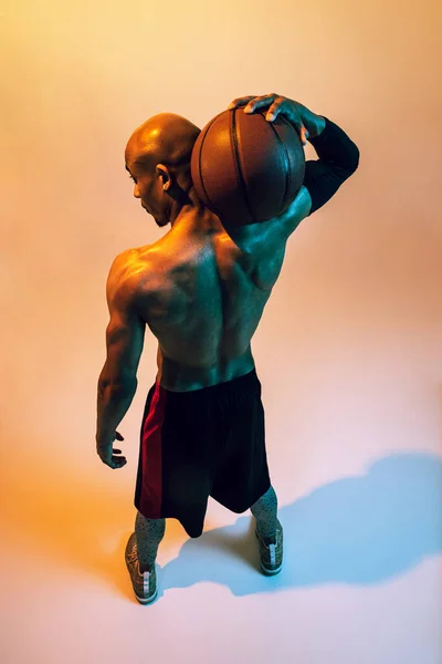 Sportive muscular african-american male basketball player posing in neon light on orange background. Back view — Stockfoto