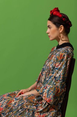 Portrait of young beautiful woman in image of famous painter, artist Frida Kahlo on green background. Side view