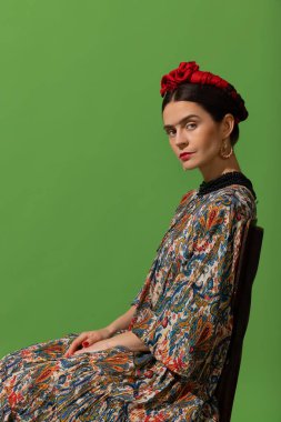 Portrait of young beautiful woman in image of famous painter, artist Frida Kahlo on green background.