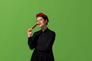 Young beautiful woman in image of famous painter, artist Frida Kahlo on green background. Retro style, comparison of eras concept.