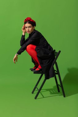 Young beautiful woman in image of famous painter, artist Frida Kahlo on green background.