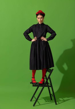 Young beautiful woman in image of famous painter, artist Frida Kahlo on green background.