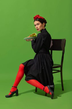 Young woman in art action remaking greatest paintings on green background. Retro style, comparison of eras concept.