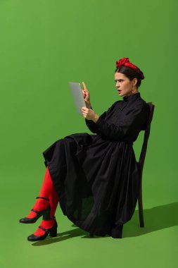 Young beautiful woman in image of famous painter, artist Frida Kahlo on green background.