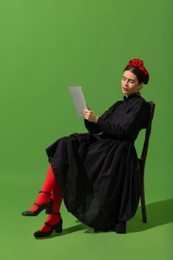 Young beautiful woman in image of famous painter, artist Frida Kahlo posing isolated over green background.