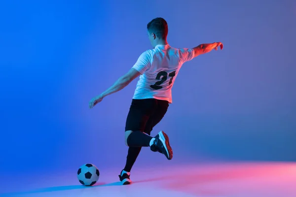 Jonge blanke man, mannelijke voetbalspeler training geïsoleerd op gradiënt blauw roze achtergrond in neon licht. Achteraanzicht — Stockfoto