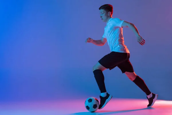 Jonge blanke man, mannelijke voetbal speler training geïsoleerd op gradiënt blauw roze achtergrond in neon licht — Stockfoto