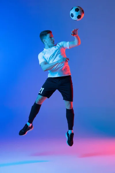 Jonge blanke man, mannelijke voetballer springen geïsoleerd op gradiënt blauw roze achtergrond in neon licht — Stockfoto