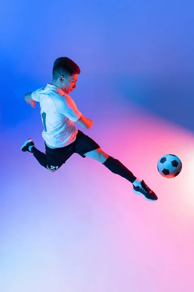 Hoge hoek uitzicht op jonge man, mannelijke voetbal speler training geïsoleerd op gradiënt blauw roze achtergrond in neon licht. — Stockfoto