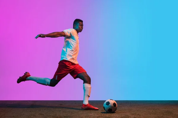 Young African man, professional soccer player playing football on grass flooring isolated on gradient blue pink background — Stock Photo, Image