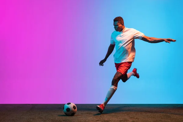 Um homem africano, treinamento de jogador de futebol profissional isolado em gradiente azul rosa fundo em luz de néon. — Fotografia de Stock