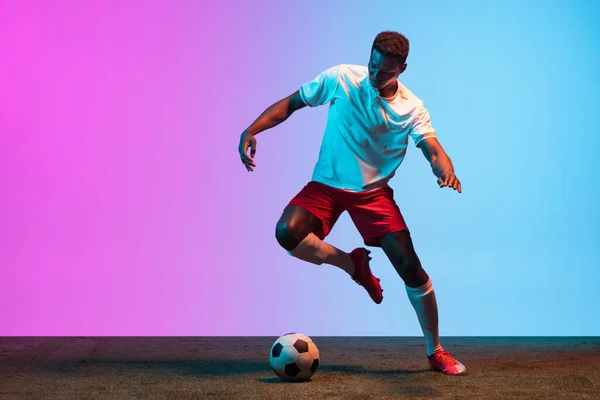 One African man, professional soccer football player training isolated on gradient blue pink background in neon light.