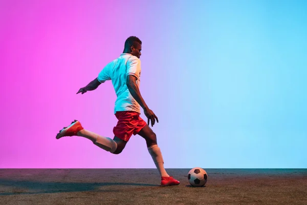 Young African man, professional soccer player playing football on grass flooring isolated on gradient blue pink background — Stock Photo, Image