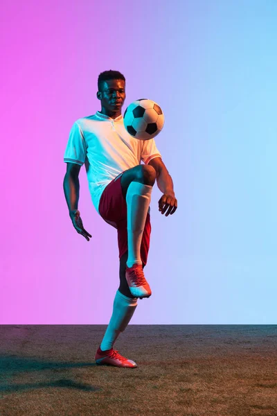 Een Afrikaanse man, professionele voetbalspeler training geïsoleerd op gradiënt blauw roze achtergrond in neon licht. — Stockfoto