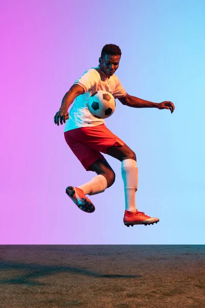 One African man, professional soccer football player training isolated on gradient blue pink background in neon light. — Stock Photo, Image