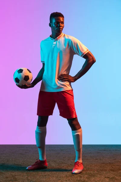 Teljes hosszúságú portré afrikai profi labdarúgó álló elszigetelt gradiens kék rózsaszín háttér neon fény. — Stock Fotó