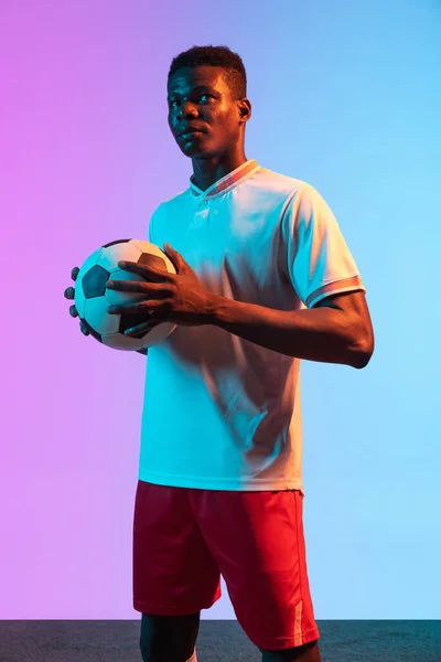 Meia-duração retrato de jogador de futebol profissional Africano de pé isolado no gradiente azul rosa fundo em luz de néon. — Fotografia de Stock
