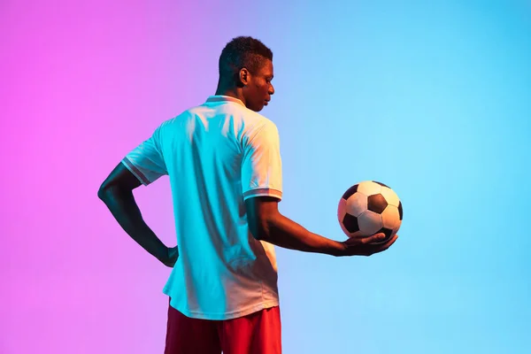 Pół-długości portret afrykańskiego profesjonalnego piłkarza stojącego na gradientowym niebieskim różowym tle w neonowym świetle. Widok z tyłu — Zdjęcie stockowe