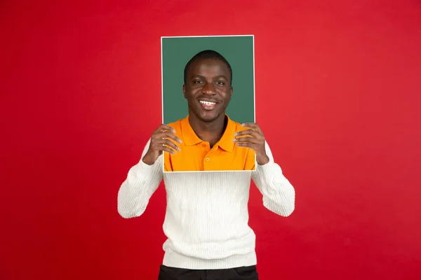 Jovem homem africano segurando seu retrato isolado sobre fundo estúdio vermelho com copyspace para anúncio. — Fotografia de Stock