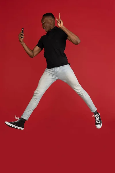 Sempre no telemóvel. Comprimento total do jovem africano tomando telefone enquanto saltava contra o fundo do estúdio vermelho. Móvel, movimento, movimento, conceitos de negócios — Fotografia de Stock