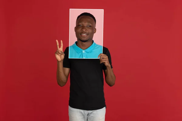 Joven hombre africano sosteniendo su retrato aislado sobre fondo rojo estudio con copyspace para anuncio. — Foto de Stock