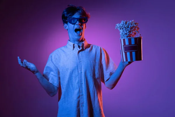 Spännande i 3D glasögon och med popcorn. Vit mans porträtt isolerad på blå studio bakgrund i rosa neon ljus. Vacker manlig modell. — Stockfoto