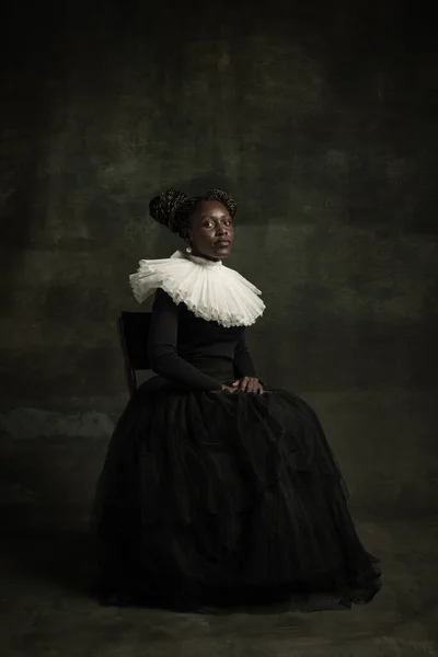 Jovem africana medieval em vestido vintage preto com grande colar branco posando isolado no fundo verde escuro. Conceito de comparação de eras, modernidade e renascimento. — Fotografia de Stock